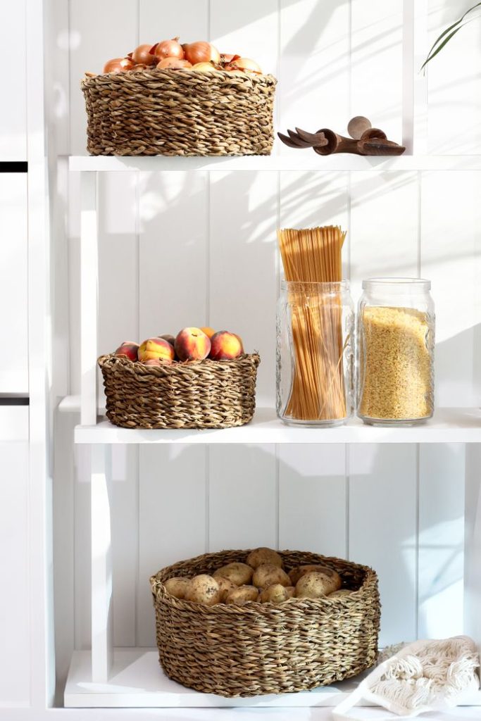 A neatly organized pantry shelf with wicker baskets holding apples, peaches, and potatoes, alongside pasta and grains.